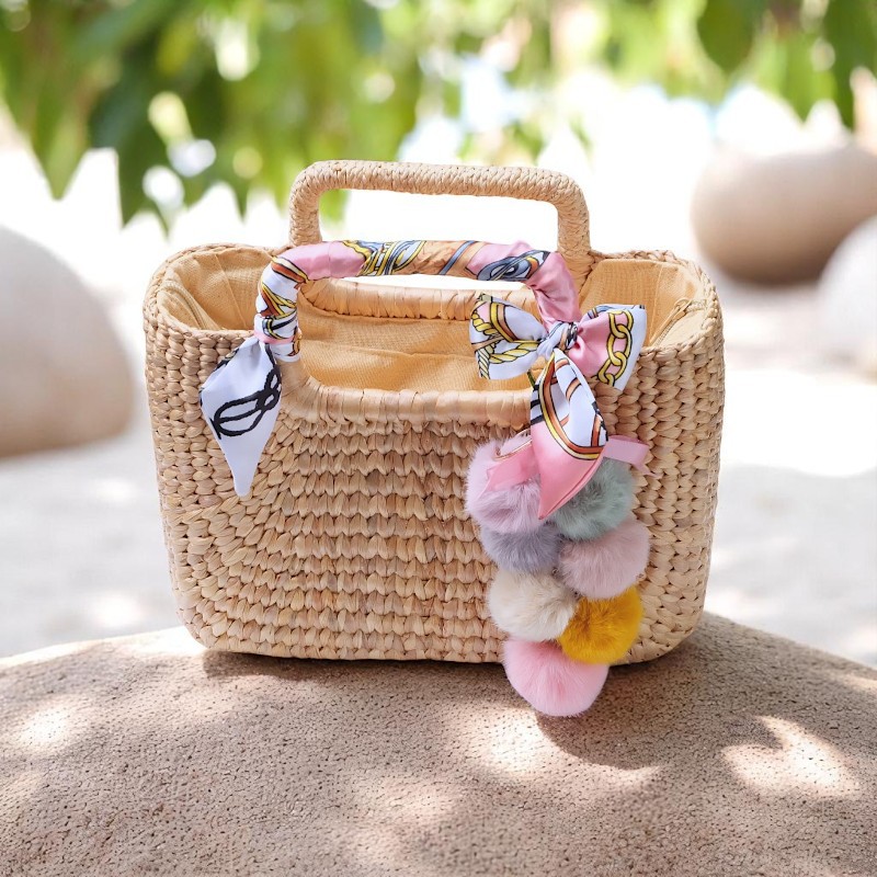 Square straw purse with pastel pom poms and scarf-wrapped handles sitting on a beachstone background